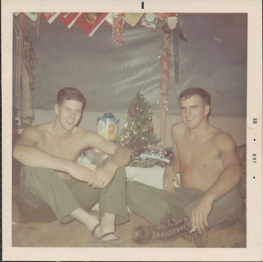 two shirtless men sit on the floor in front of a small decorated christmas tree, smiling at the camera. The photo has a…