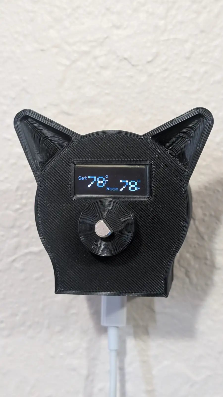 A black 3D-printed cat-shaped thermostat with cat ears and a digital display showing 'Set 78°F' and 'Room 78°F', plugged…