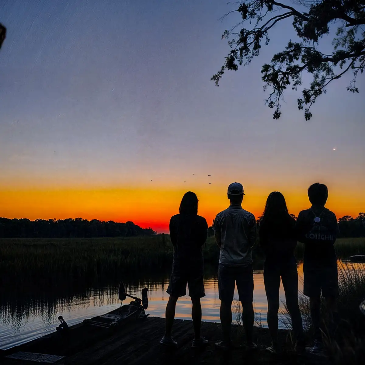 Four people stand in silhouette on a dock at sunset, watching the orange and purple sky over water with birds flying. The…
