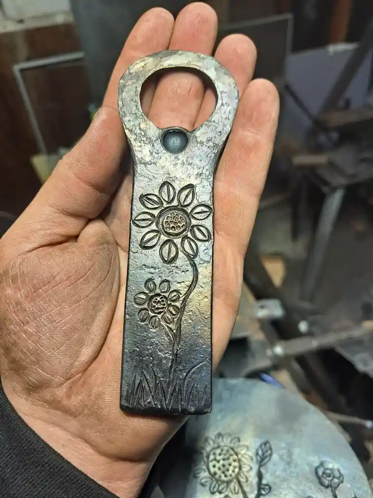 A hand holds a handmade, rustic metal bottle opener with an engraved sunflower and smaller flower design. The opener has a…