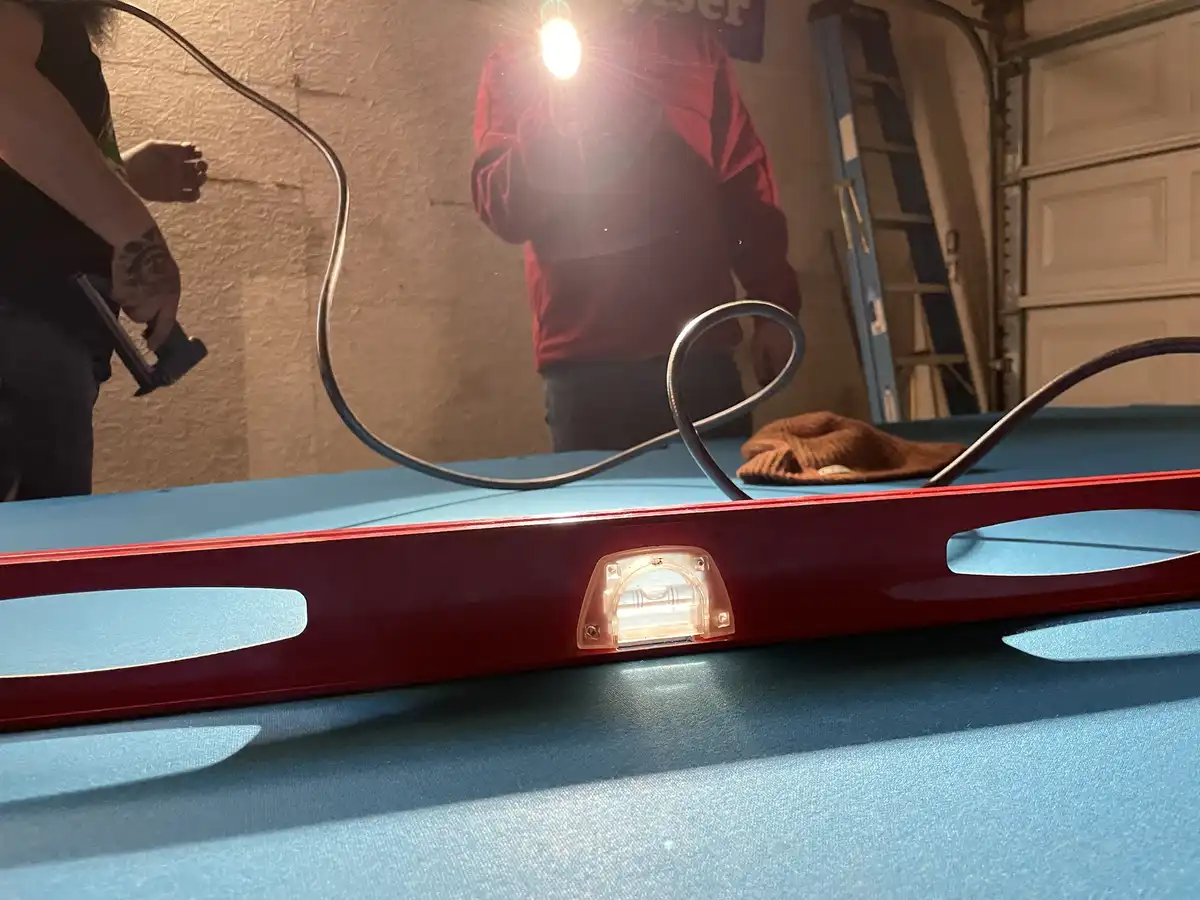 A close-up shot of a red spirit level resting on a blue pool table, with a bright light source causing lens flare. In the…