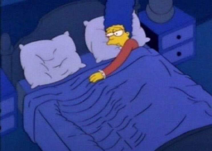 marge simpson lies in bed under a blue blanket, looking visibly annoyed or frustrated, with her eyes narrowed and one hand…