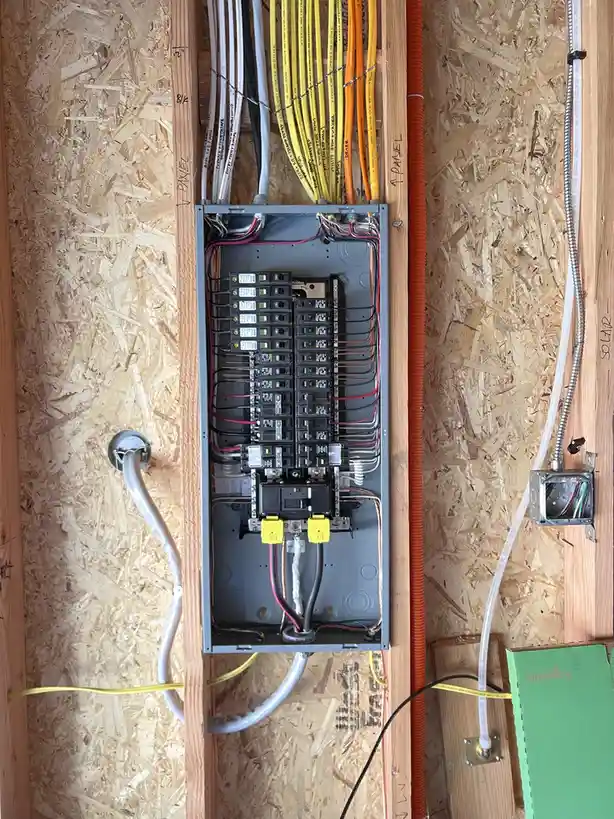 An overhead view of an open electrical panel mounted on oriented strand board (OSB) walls, with numerous wires of various…