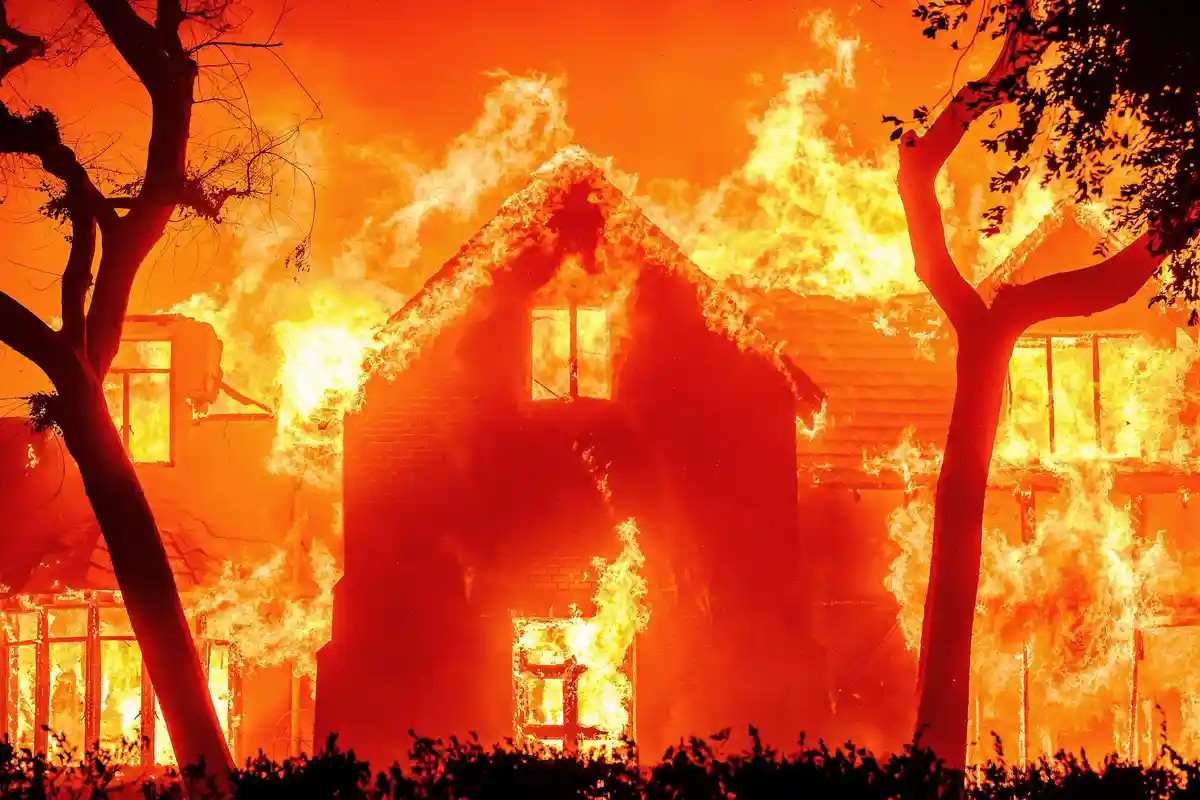 A house is engulfed in bright orange and yellow flames, with fire consuming its roof, windows, and walls, framed by…