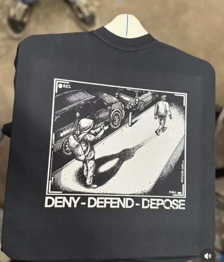 A black t-shirt displays a black-and-white graphic resembling a security camera feed showing a person in a hoodie filming…