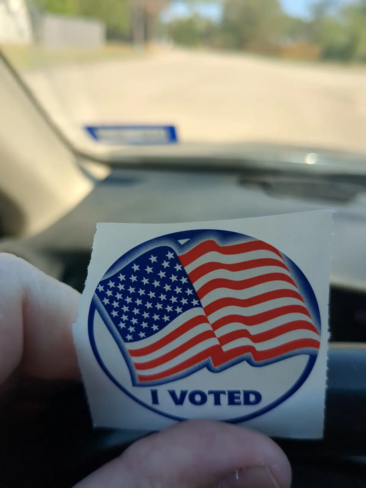 A hand holds up a circular 'I VOTED' sticker featuring a waving American flag design, taken from inside a car with the…