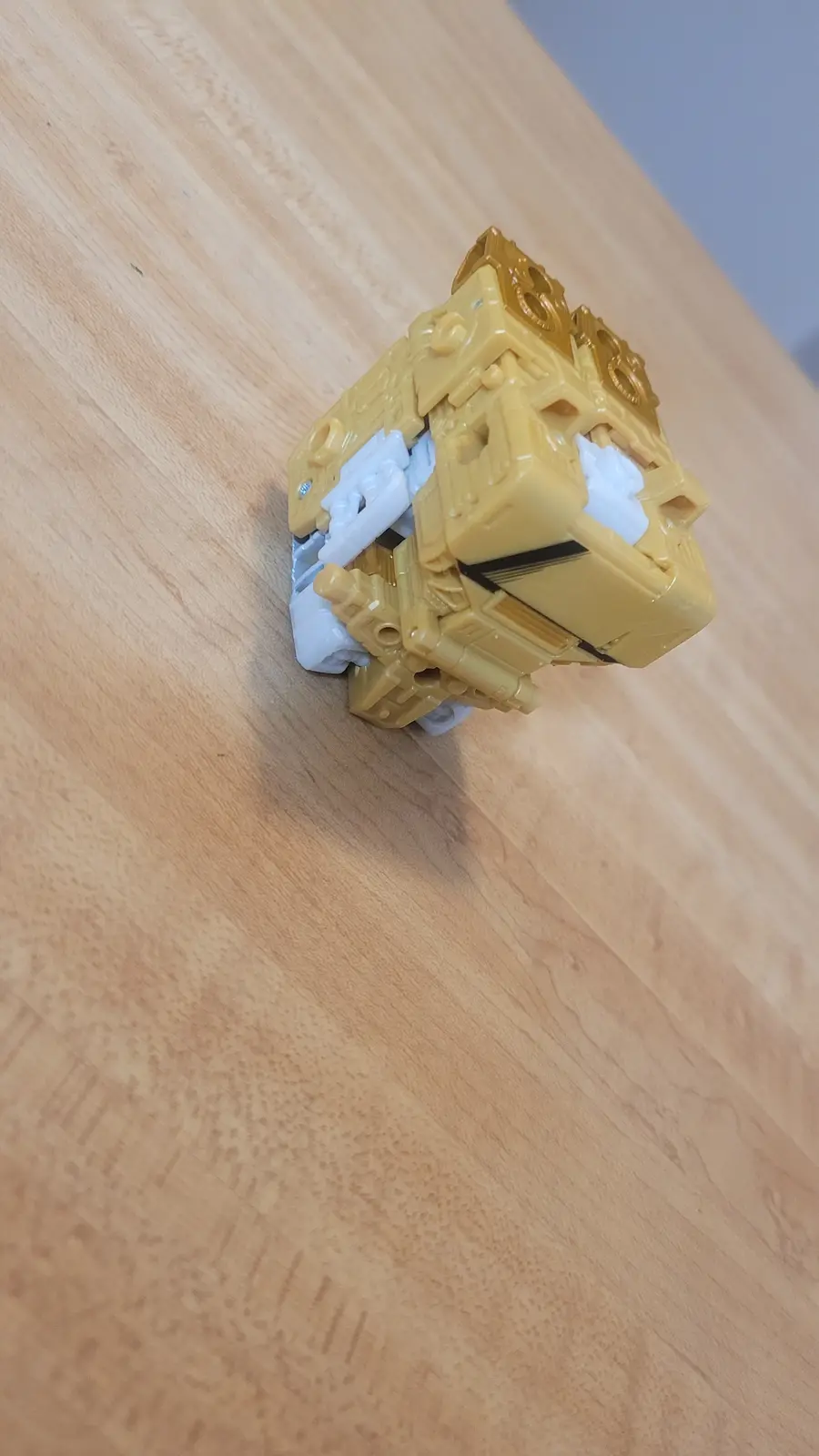 A yellow and white plastic toy robot, resembling a Transformer, is shown in a compact, cube-like form on a light wooden…