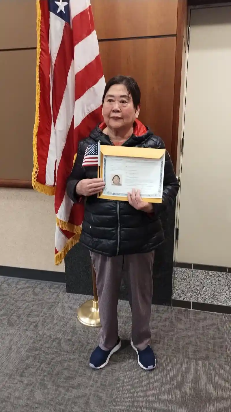 An older woman stands indoors in front of an American flag, holding a framed citizenship certificate in one hand and a small…