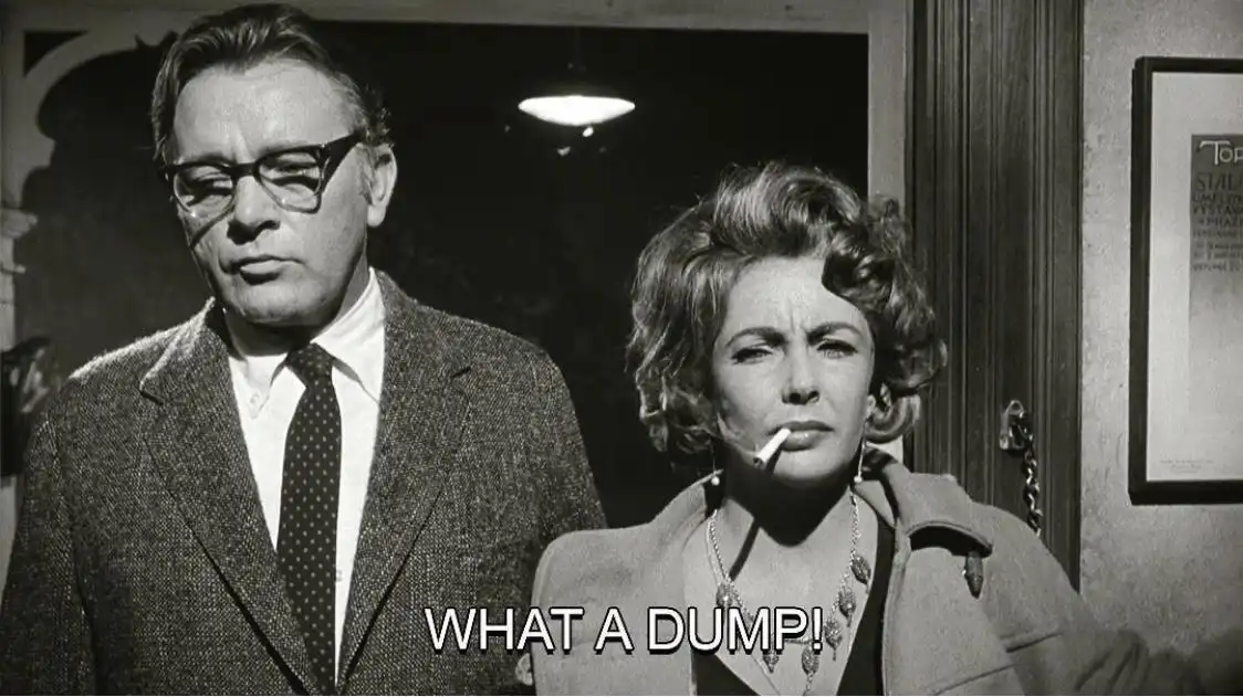 A black-and-white still from a vintage film shows a man in glasses and a suit standing beside a woman with styled hair…