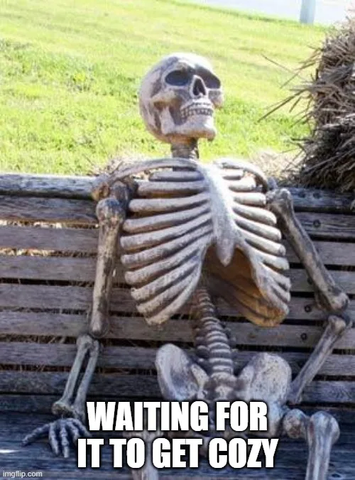 A plastic skeleton sits relaxed on a wooden park bench with grass and hay bales in the background. The skeleton is posed…