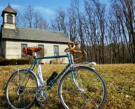 A teal road bicycle with wooden handlebars and a brown leather saddle is parked on grass in front of a small white church…