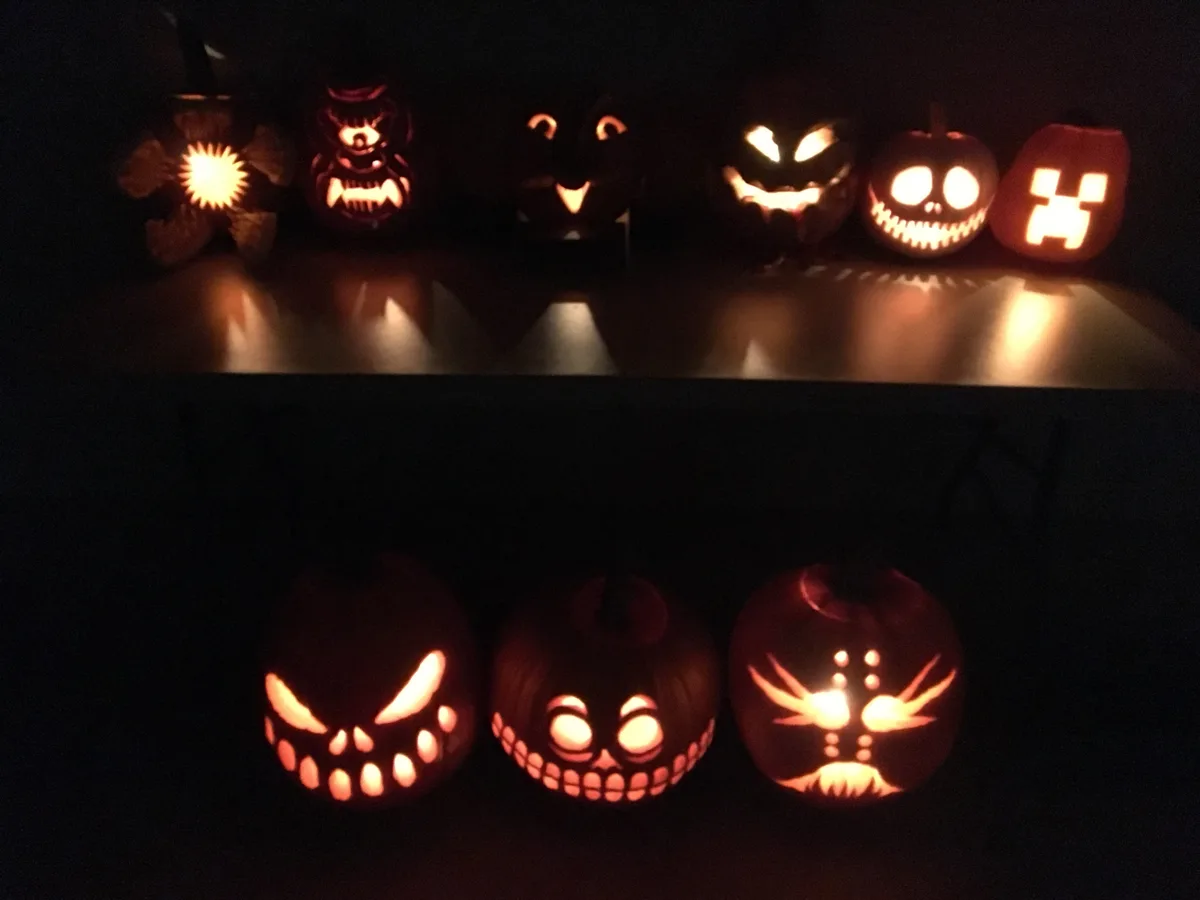 A dimly lit photo shows two rows of carved pumpkins glowing from within. The top row features six pumpkins with various…