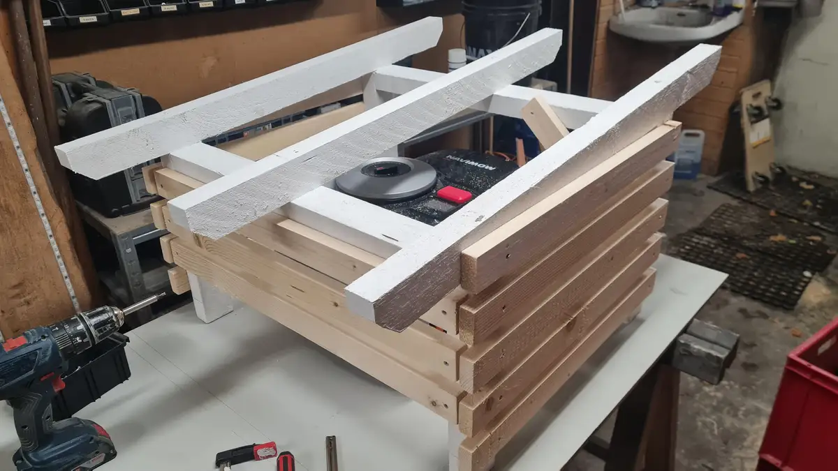 A wooden box with white-painted beams is being constructed on a workbench, with a robot vacuum cleaner (brand Navimow)…