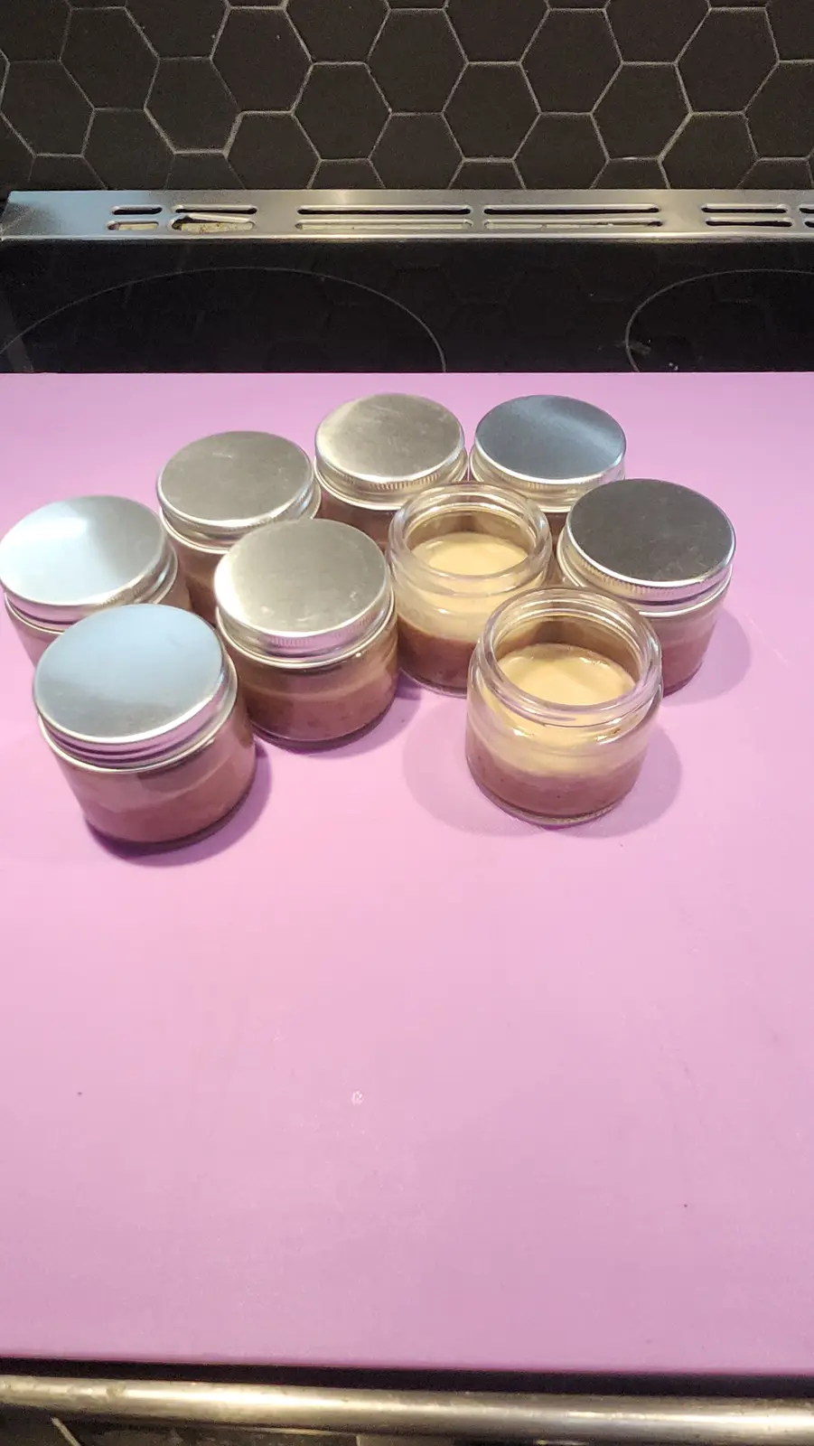 A photo shows nine small glass jars with silver lids arranged on a pink countertop, in front of a black hexagonal tile…