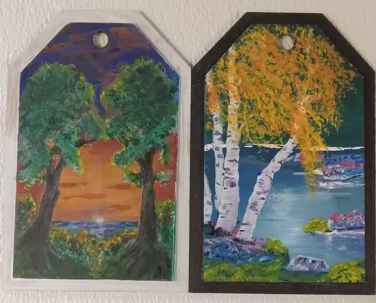 Two hand-painted, octagonal wooden tags hang side by side against a textured white wall. The left tag depicts two green…