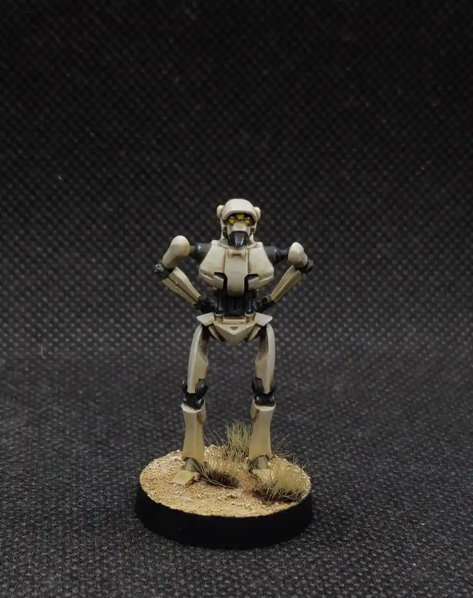 A painted miniature of a beige and black humanoid robot stands on a circular base with sandy terrain and sparse grass, arms…