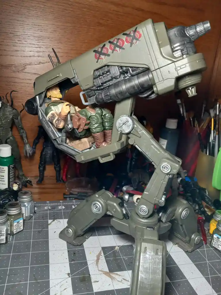 A custom-painted green mech toy with a bearded action figure pilot sits on a gridded work mat surrounded by paint supplies…