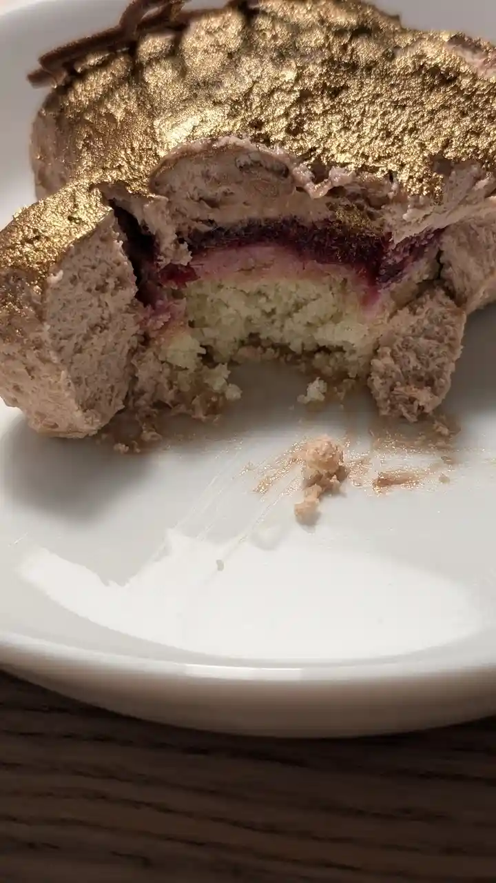 A close-up photo shows a partially eaten dessert on a white plate, revealing layers of cake, cream, and jam beneath a shiny…