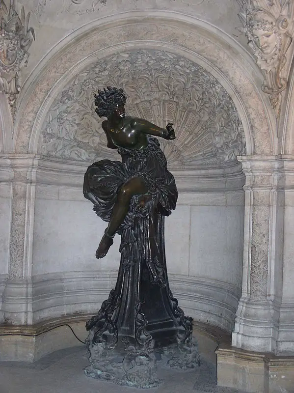 A bronze statue of a woman in a flowing dress, posed dynamically as if dancing or moving, stands inside an ornate stone…