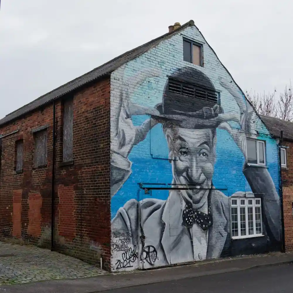 A large mural on the side of a brick building depicts a smiling man in a bowler hat and bow tie, with his hands adjusting…