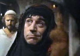 A man in a dark head covering stares with a confused and slightly alarmed expression. Behind him, another man in a white…