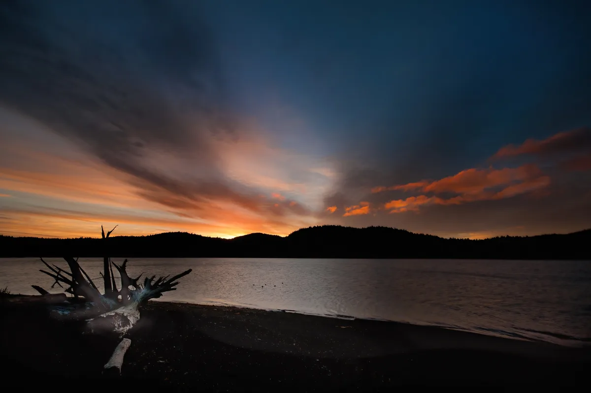 A serene sunset over a calm lake with dark silhouetted hills and a large piece of driftwood on the shore. The sky is painted…