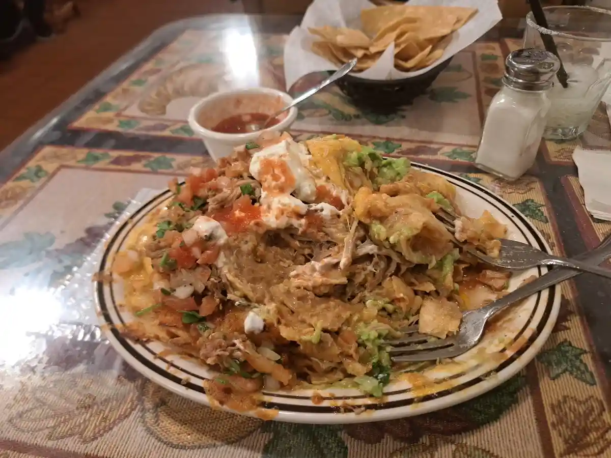 A partially eaten plate of cheesy, saucy Mexican food, likely tacos or nachos, sits on a patterned tablecloth with forks…
