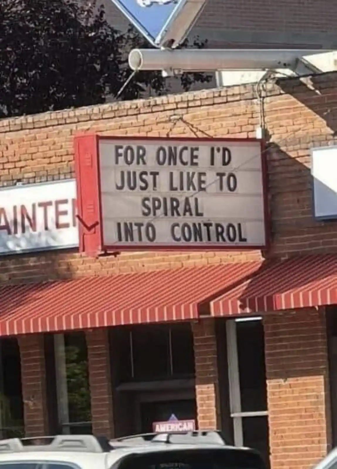A red-framed marquee sign on a brick building reads 'FOR ONCE I'D JUST LIKE TO SPIRAL INTO CONTROL'. The sign is mounted…