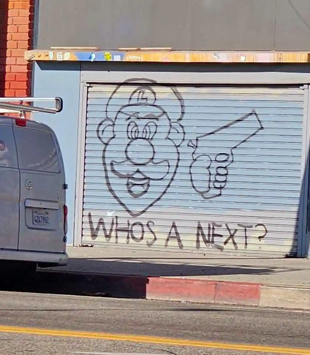 A crude black outline graffiti on a metal roll-up door depicts a cartoonish mustachioed man with a cap holding a pistol…