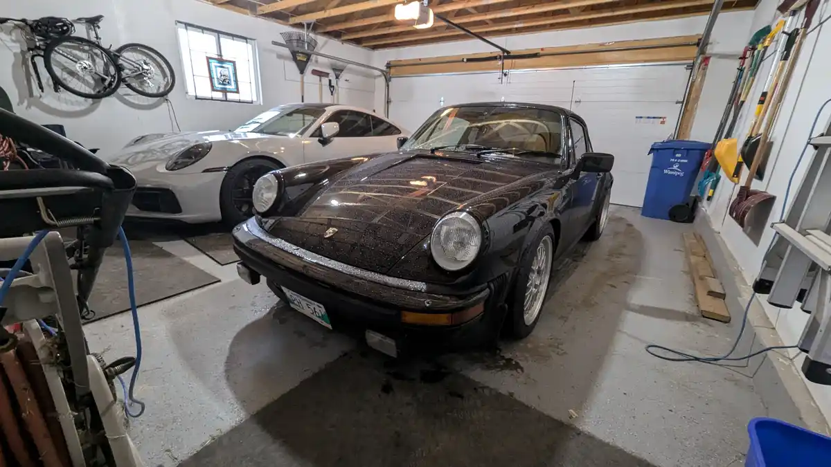 A garage contains a black classic Porsche 911 Turbo in the foreground, visibly wet with water droplets, and a modern white…