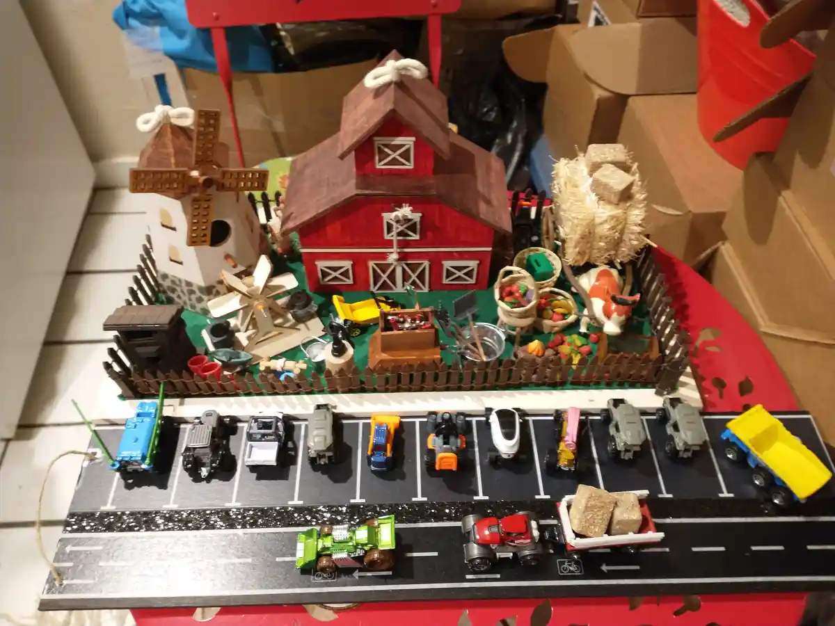 A handmade toy farm diorama featuring a red barn, windmill, fences, hay bales, and a plastic cow, sits behind a parking lot…