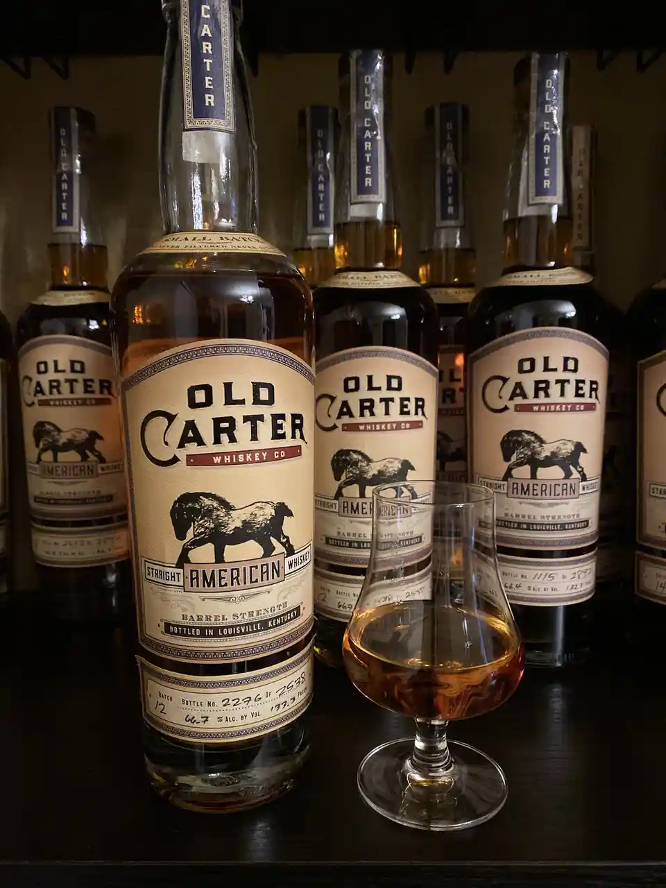 A close-up photo shows multiple bottles of Old Carter Whiskey Co. arranged on a shelf, with one bottle prominently in the…