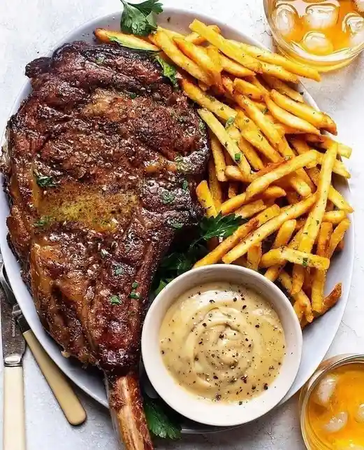 A top-down photo of a large, seared tomahawk steak with a long bone, served on a white plate alongside golden french fries…