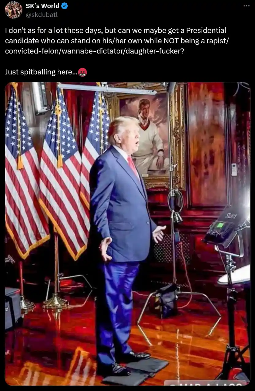 A man in a blue suit stands on a stage with American flags behind him, gesturing as if speaking. A framed portrait of a man…