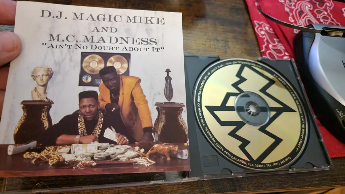 A hand holds a CD case for 'D.J. Magic Mike and M.C. Madness' album 'Ain't No Doubt About It', showing two men in gold…