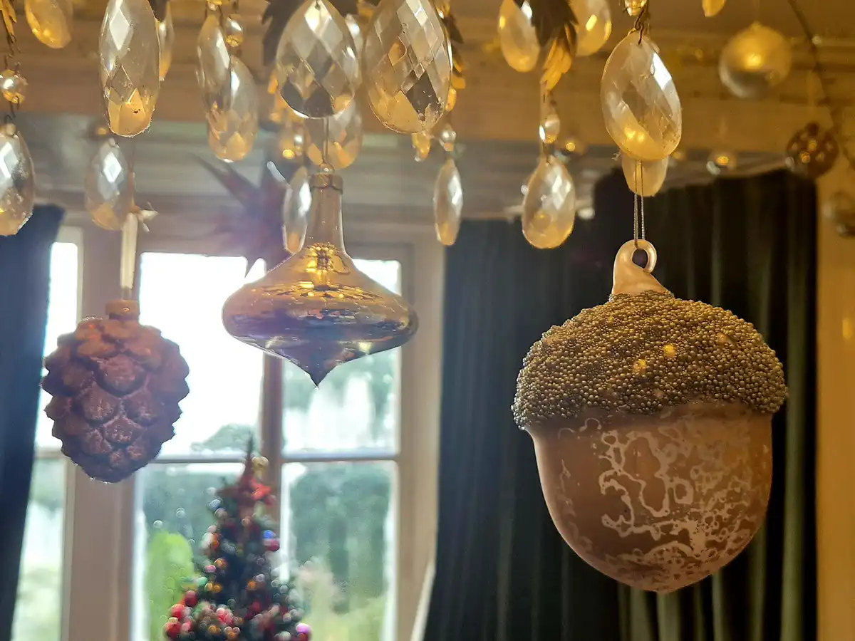 A close-up photo shows several ornamental Christmas decorations hanging from a chandelier, including a pinecone, a glass…