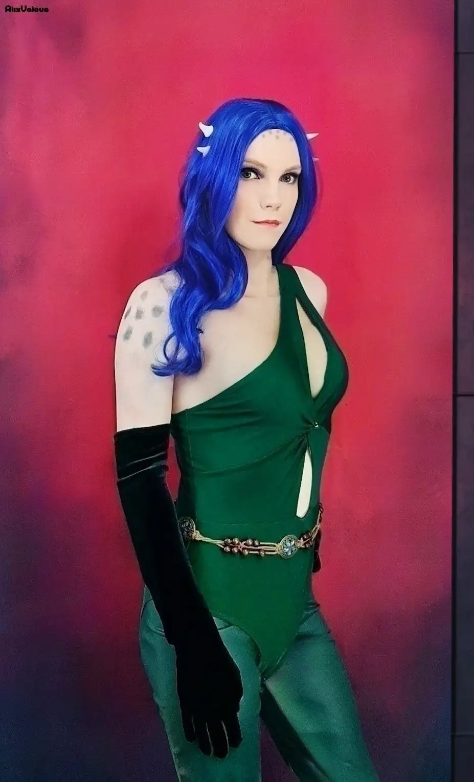 A person with long blue hair and small white horns poses in a green one-piece outfit with black gloves and a decorative belt…