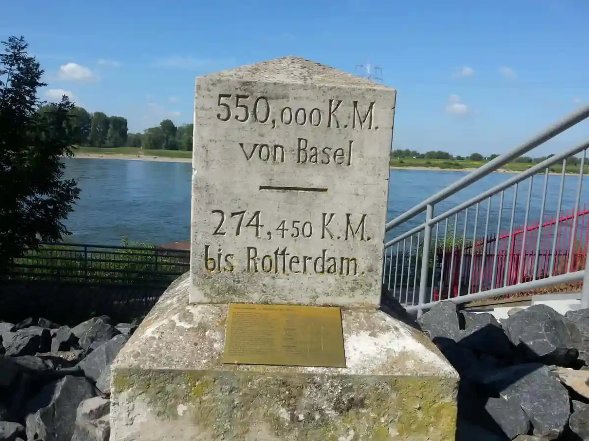 A weathered stone marker by a river shows '550,000 K.M. von Basel' above a line and '274,450 K.M. bis Rotterdam.' below…