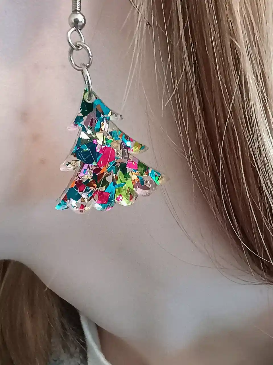 A close-up photo shows a person's ear adorned with a sparkly, multi-colored Christmas tree-shaped earring made of glittery…