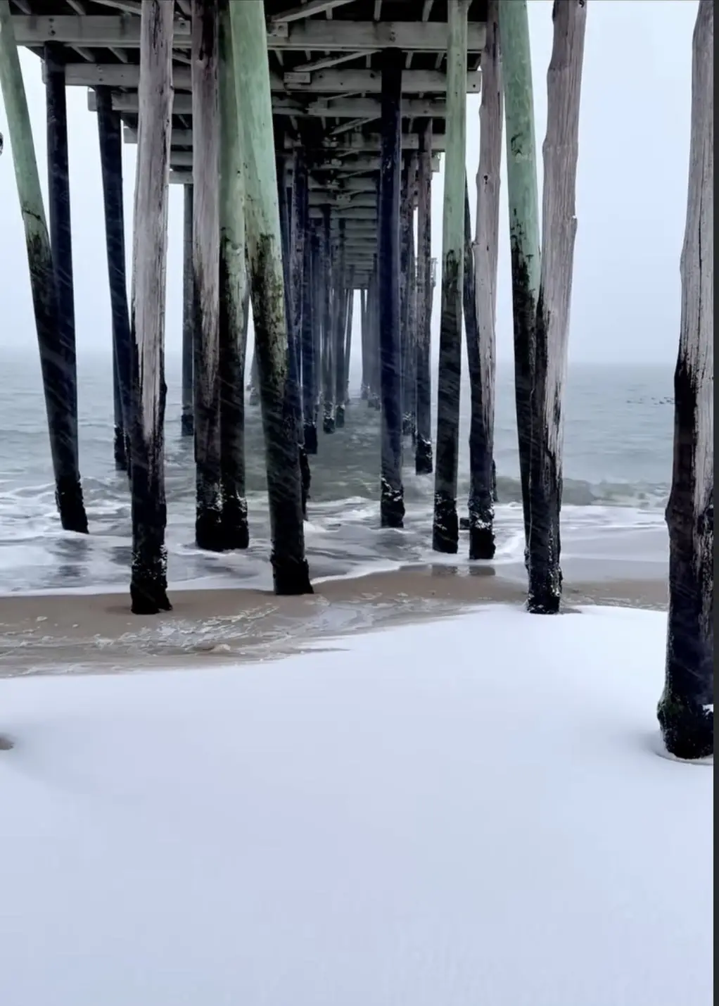 A beach scene under a pier with snow covering the sand, waves lapping at the pilings, and a foggy ocean in the background…