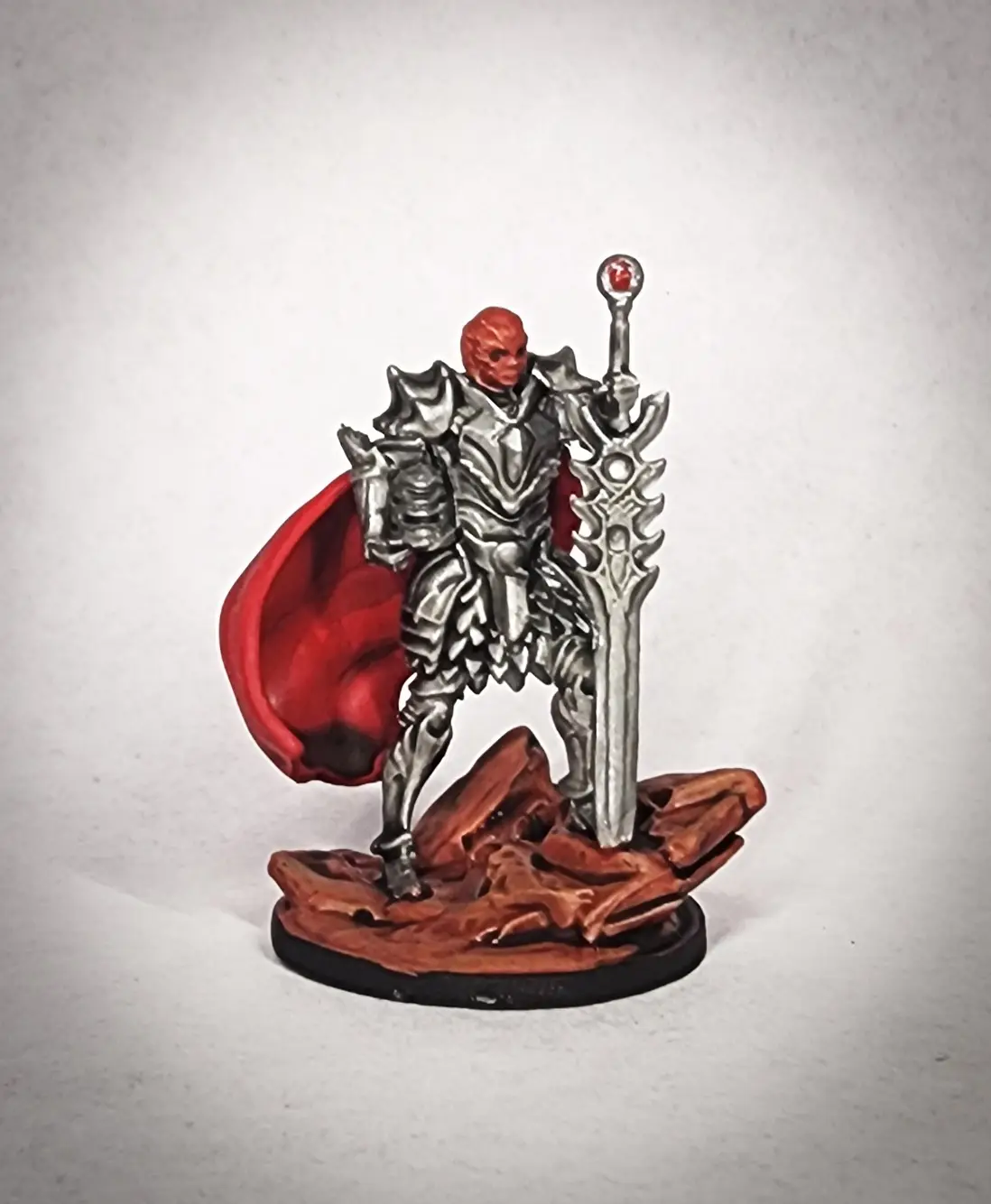A painted miniature figurine of a red-skinned, armored warrior with a red cape, standing on a rocky base and holding a large…