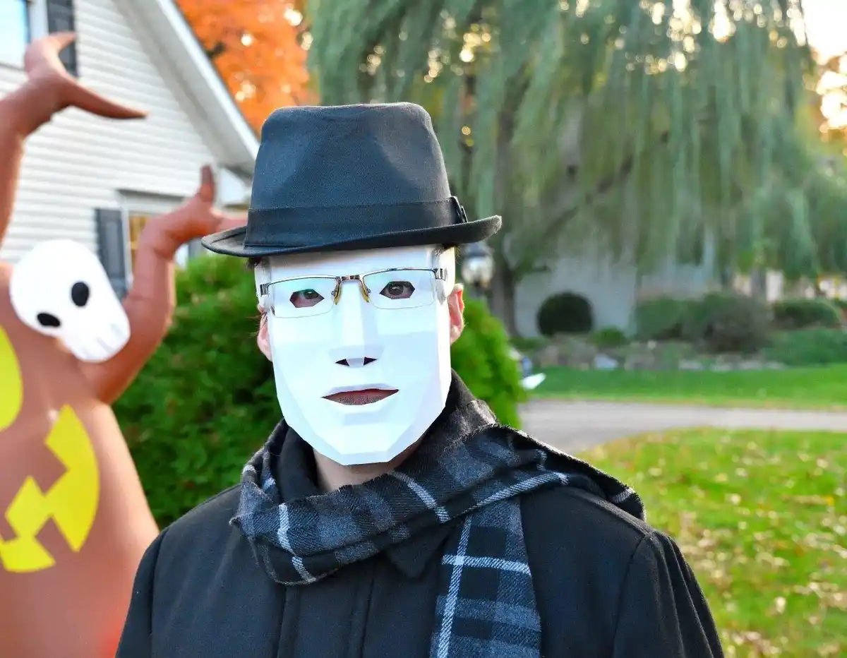 A person wearing a white geometric mask, glasses, a black fedora, and a plaid scarf stands outdoors in front of a house with…