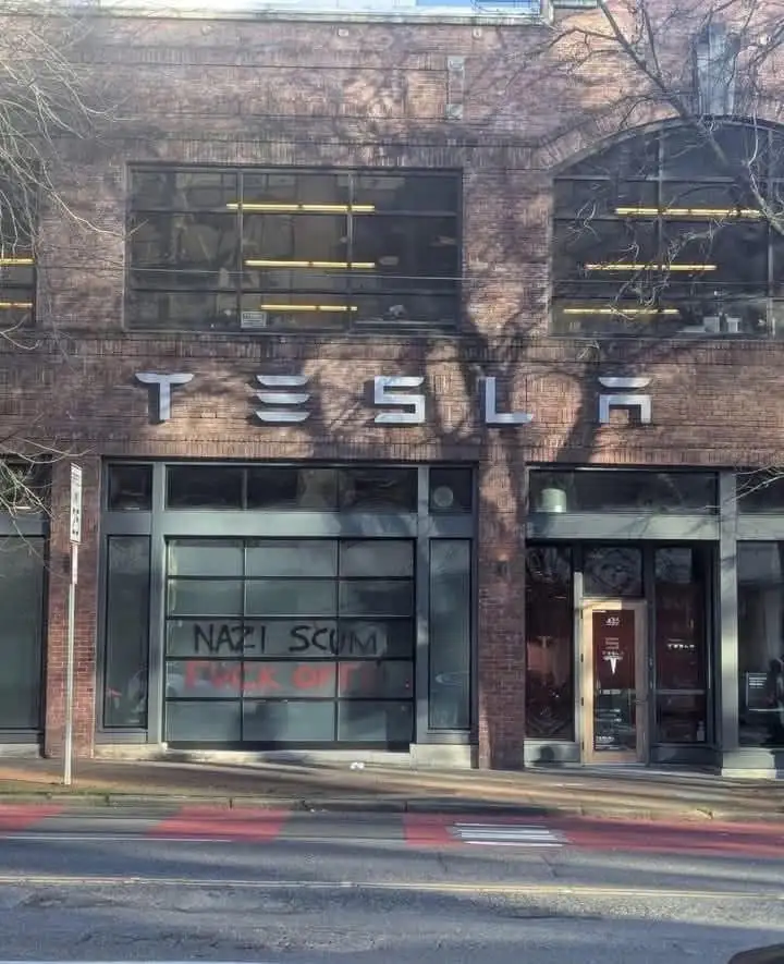 A brick building with a Tesla logo above its entrance has graffiti on the glass door reading 'NAZI SCUM FUCK OFF'. The…