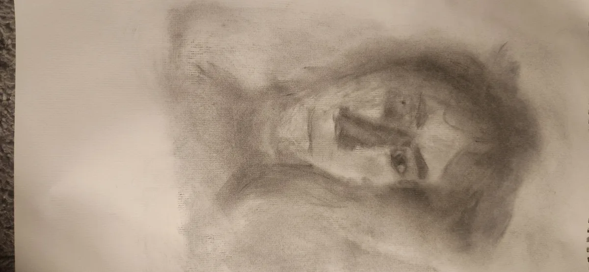 A blurry, smudged charcoal sketch on paper depicts an indistinct, abstract human-like face with vague eyes and a mouth. The…