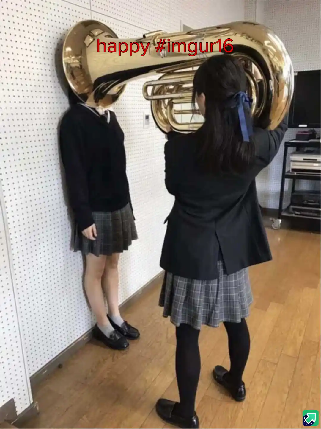 Two girls in school uniforms stand in a room; one holds a large brass tuba, positioning its bell to cover the face of the…