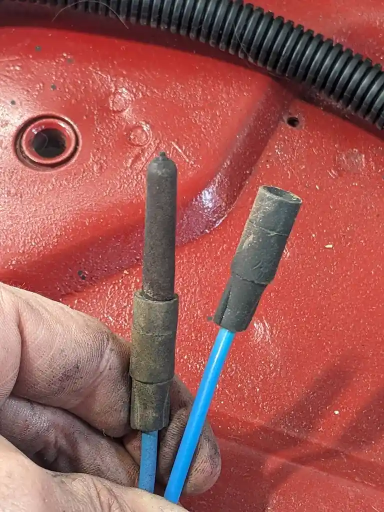 A hand holds two spark plug wires against a red engine block; one wire has a worn, dirty, and corroded terminal, while the…