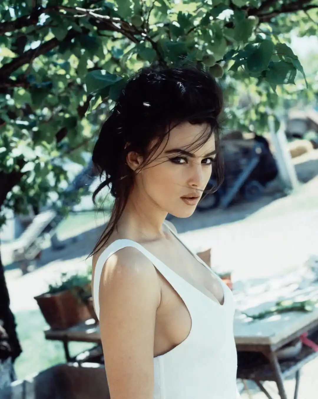 a woman with dark hair in a white top stands outdoors under green foliage, looking over her shoulder with a serious…