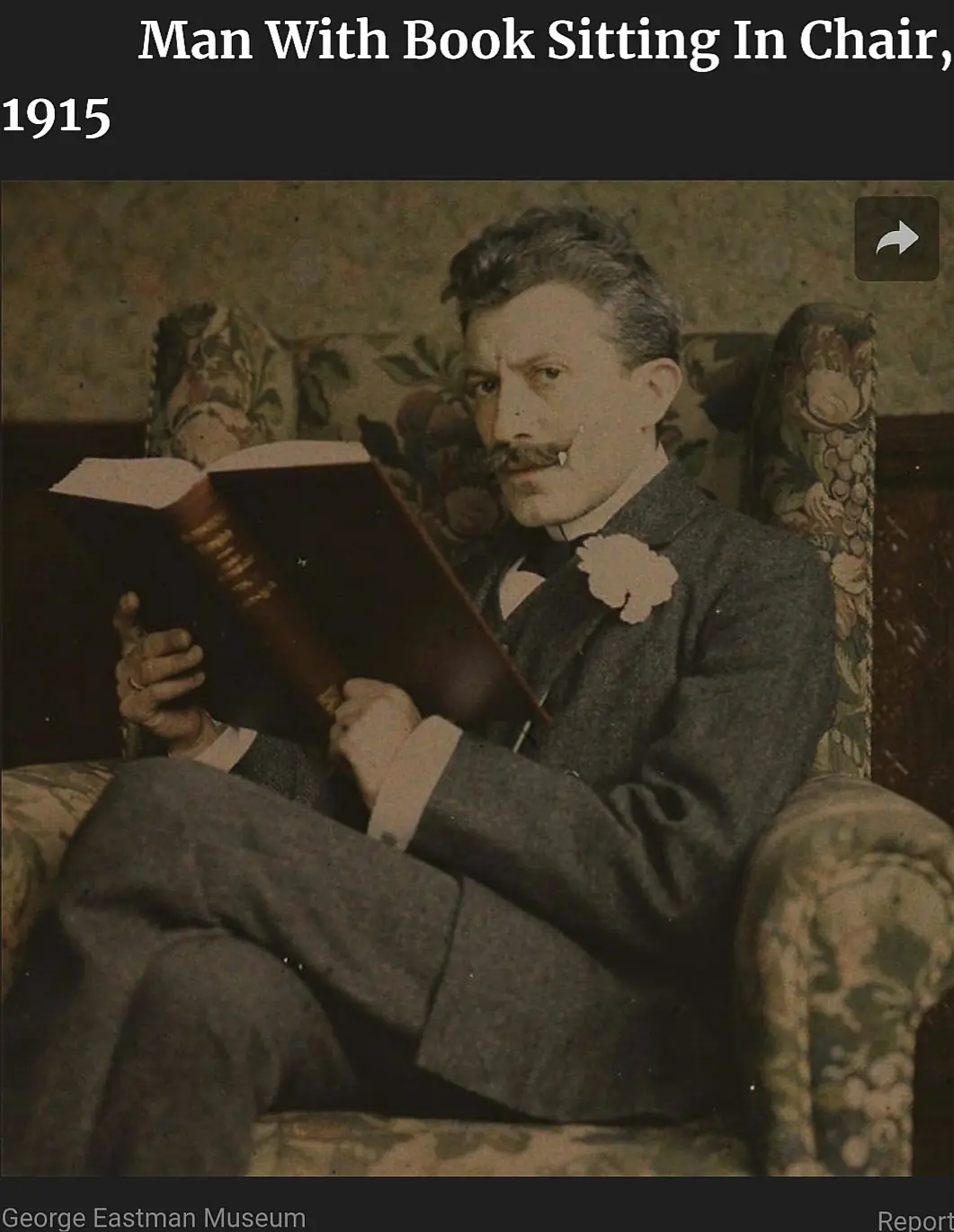 A vintage 1915 photograph shows a man in a suit with a mustache and a flower boutonniere, sitting in a floral-patterned…