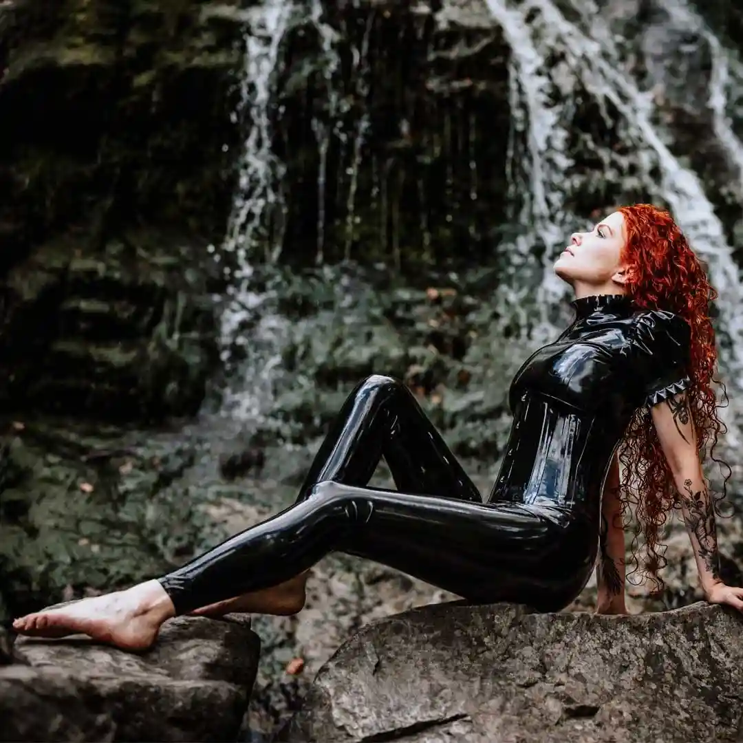 a woman with long red hair and tattoos sits barefoot on rocks in front of a waterfall, wearing a shiny black latex bodysuit…