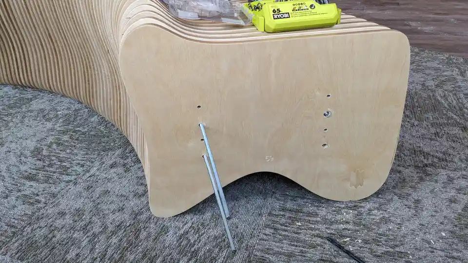 A close-up of the underside of a curved wooden bench under construction, showing a metal rod inserted into a hole, with a…